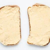 Top view photo of hummus spread on top of two slices of toast. The toast is on a white tabletop.