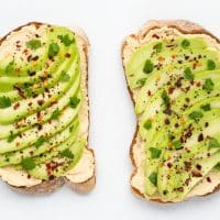 Top view photo of hummus toast, topped with slices of avocado, black pepper, red pepper flakes, and cilantro. The toast is on a white tabletop.