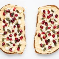 Top view photo of hummus toast topped with pomegranate seeds and pumpkin seeds. The toast is on top of a white tabletop.