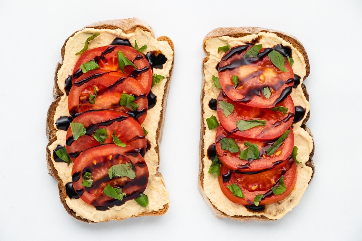 Top view photo of two slices of hummus toast, topped with tomato slices, drizzled with balsamic glaze, and sprinkled with fresh basil. The toast is sitting on a white tabletop.