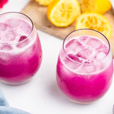 Photo of two small glasses, filled with Dragon Fruit Lemonade and ice. In the background are lemon halves that have been juiced and a wooden cutting board.
