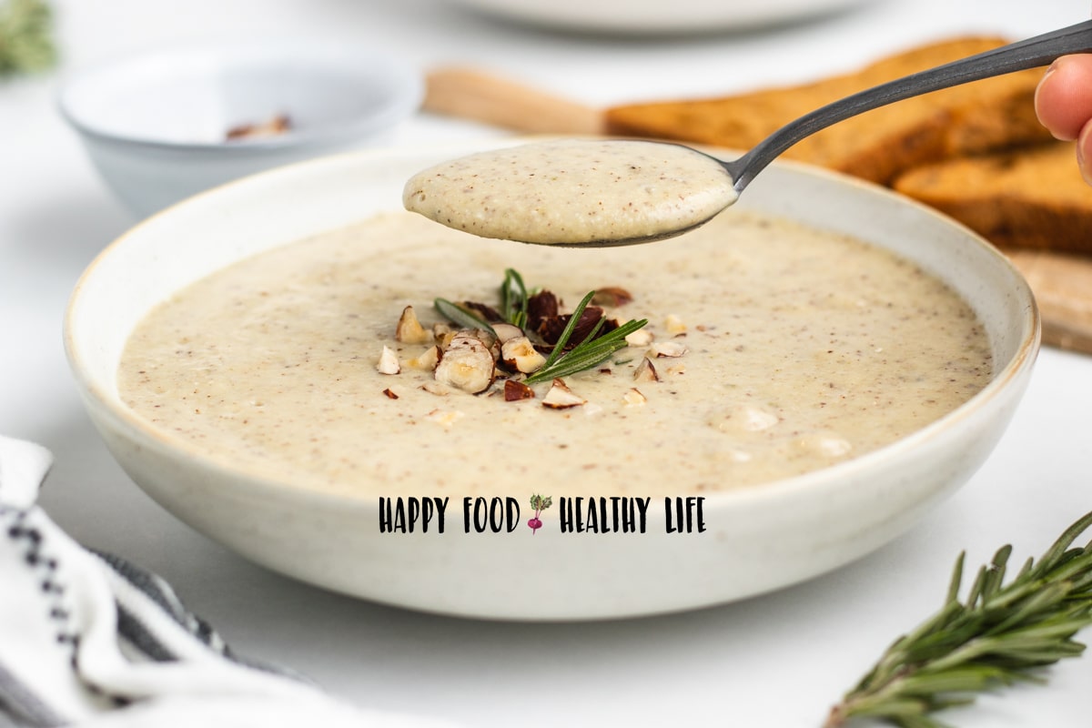 Photo of a white bowl of hazelnut soup. It is topped with fresh rosemary and chopped hazelnuts. There is a silver spoon hovering above the bowl, with a spoonful of soup in it. In the foreground is fresh rosemary and a kitchen towel. In the background is bread and another bowl of soup. 