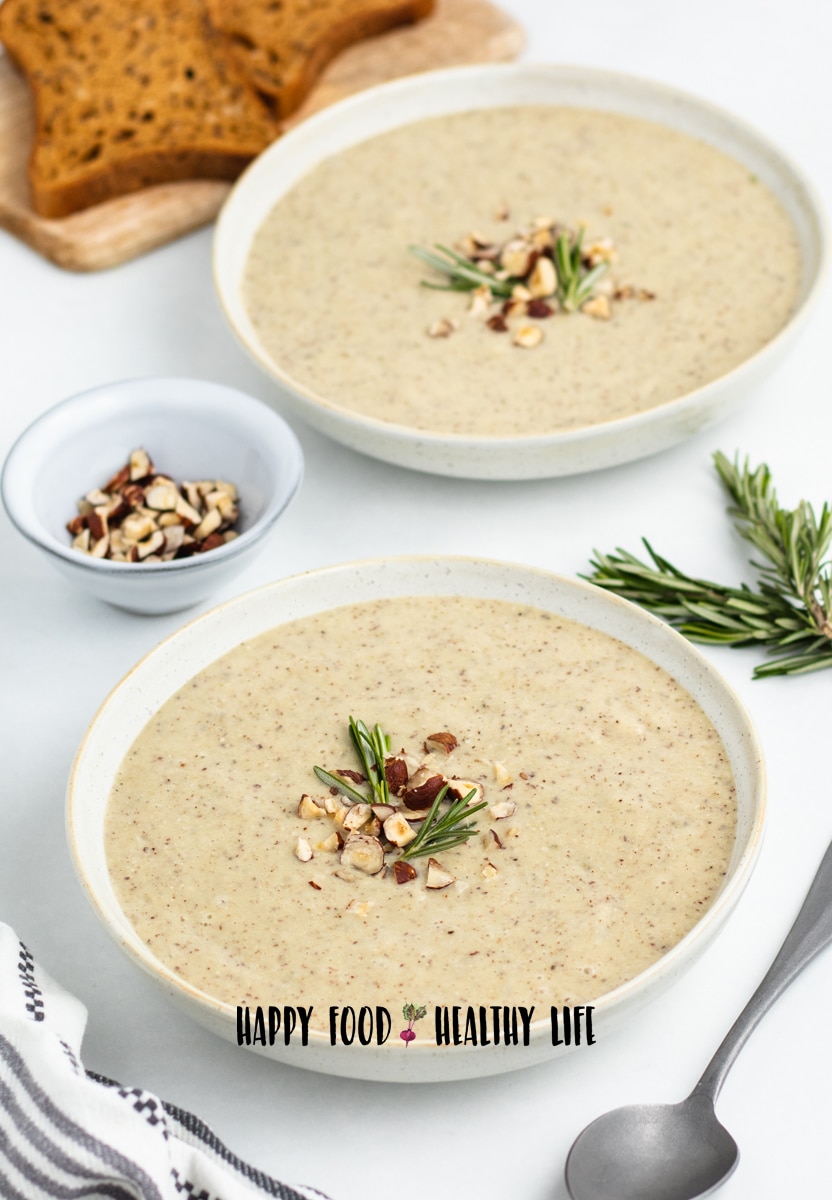 Photo of  two white bowls of hazelnut soup. They are both topped with chopped hazelnuts and fresh rosemary. There is a silver spoon in the foreground and bread in the background. 