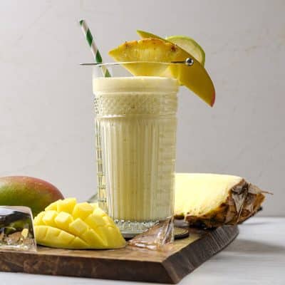 Photo of a glass, filled with Pineapple Mango Smoothie. There is a pineapple, mango, and lime garnish on the glass rim, and a striped green straw in the cup. The cup is on a wooden cutting board and there is a mango half in the foreground. There is a pineapple wedge in the background.