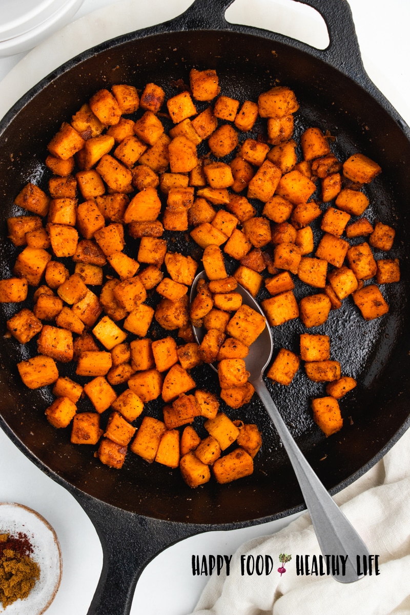 Top view photo of a cast iron skillet with sauteed sweet potato cubes in the pan. There is a silver in the pan, and a small bowl with spices in the bottom of the photo. 