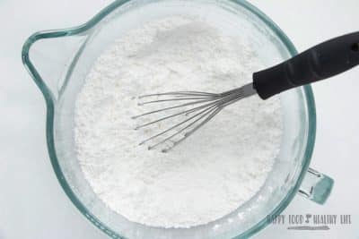 a clear glass mixing bowl with a spout, filled with powdered sugar. A black handled whisk is in the bowl, and it's viewed from overhead. Photo Credit: Cindy Gordon of HappyFoodHealthyLife.com