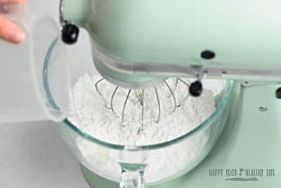 a pastel green stand mixer fitted with a whisk attachment and a clear glass bowl. In the bowl is powdered sugar. Water is being poured in with a measuring cup. Photo Credit: Cindy Gordon of HappyFoodHealthyLife.com