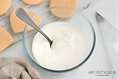 closeup shot of a clear glass bowl of white creamy vegan royal icing with a small spoon it in. To the right is an empty plastic piping bag fitted with a small round tip. To the left is a tan linen napkin. In the top left corner is a wire rack of undecorated heat and circle shaped cookies. Photo Credit: Cindy Gordon of HappyFoodHealthyLife.com