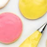 closeup of two round cut out cookies decorated with vegan royal icing one is pink, the other is yellow. Next to them is a piping bag fitted with a #2 tip, filled with yellow frosting. Photo Credit: Cindy Gordon of HappyFoodHealthyLife.com